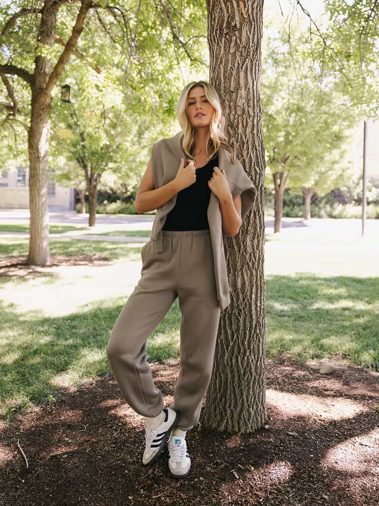 A woman with long blonde hair leans against a tree in a park, dressed casually in Cozy Earth's Women's CityScape Crewneck paired with a beige cardigan and matching beige joggers. She completes her outfit with white sneakers, standing amidst greenery and sunlight filtering through the trees. |Color:Ebony