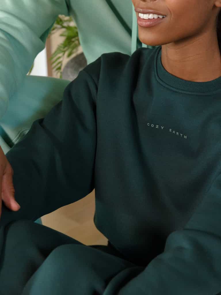 A person wearing a dark green Women's CityScape Crewneck from Cozy Earth is sitting indoors. The image focuses on the upper body and part of the arm, with a soft smile visible. A blurred plant is in the background. |Color:Spruce