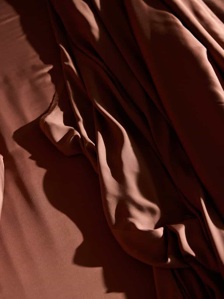 The Bamboo Flat Sheet - Wholesale by Cozy Earth exhibits a rich, brown satin appearance, artfully draped and softly illuminated by gentle light, creating smooth folds and shadows. The silky, luxurious texture of the material reflects light to emphasize its sheen and fluidity. |Color:Terra