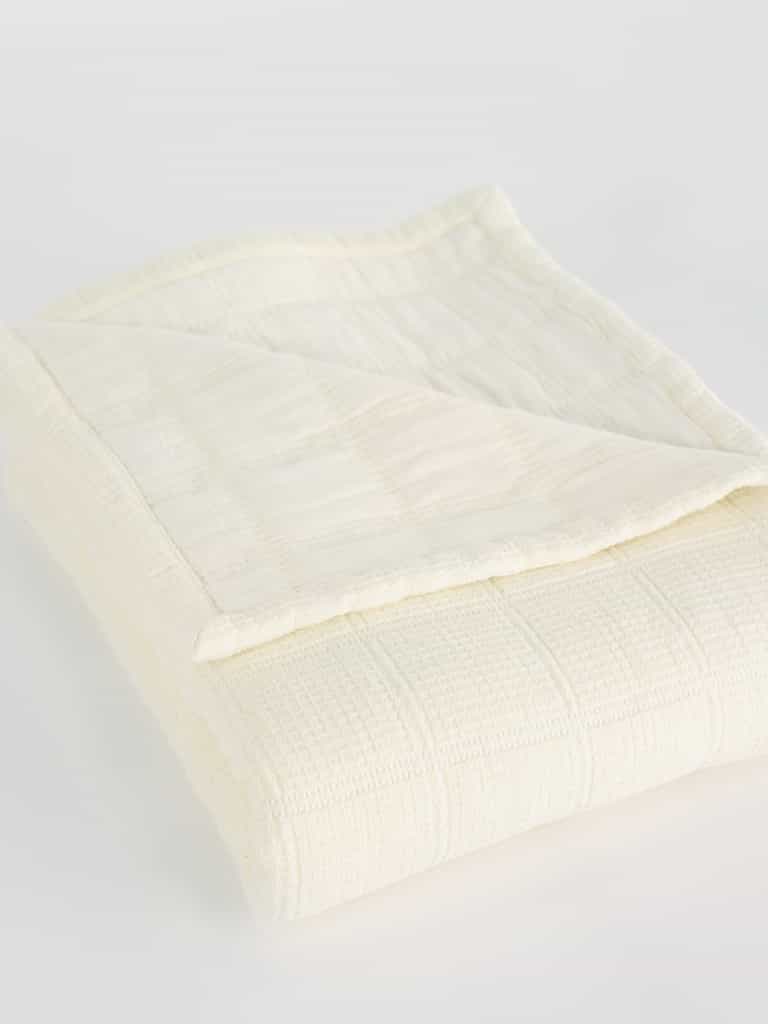A neatly folded Waffle Windowpane Blanket from Cozy Earth, cream-colored with a subtle checkered pattern. The blanket has a soft texture and looks lightweight, perfect for adding a cozy layer to a bed or couch. The corner of the blanket is flipped over, revealing its delicate stitching and fabric detail. |Color:Creme