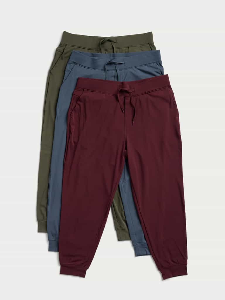 Three pairs of jogger pants are neatly arranged, overlapping each other. The colors from top to bottom are olive green, navy blue, and maroon. Each pair is designed with a drawstring waistband and cuffed ankles from Cozy Earth's Men's Ultra-Soft Bamboo Pullover Crew collection. |Color:Olive