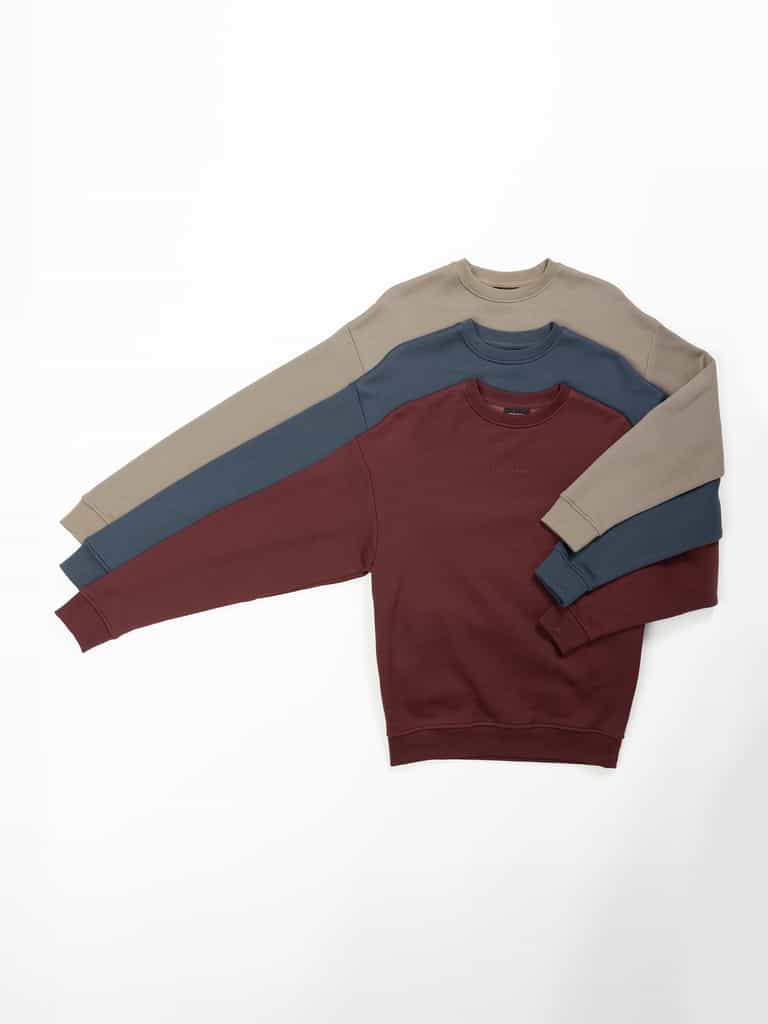 Three Women's CityScape Crewneck sweaters from Cozy Earth, available in beige, blue, and maroon, are neatly stacked on a plain white background. Each sweater features long sleeves and a simple design. |Color:Burgundy