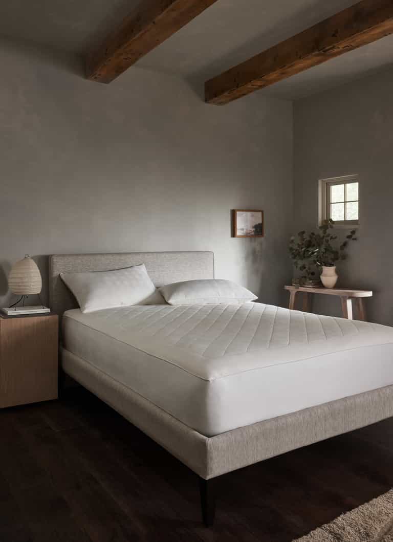 A minimalist bedroom features a light gray upholstered bed with white bedding topped by the Cozy Earth Bamboo Mattress Pad, wooden ceiling beams, dark wood flooring, a small nightstand with lamp, a table with greenery, and a small window on the wall.