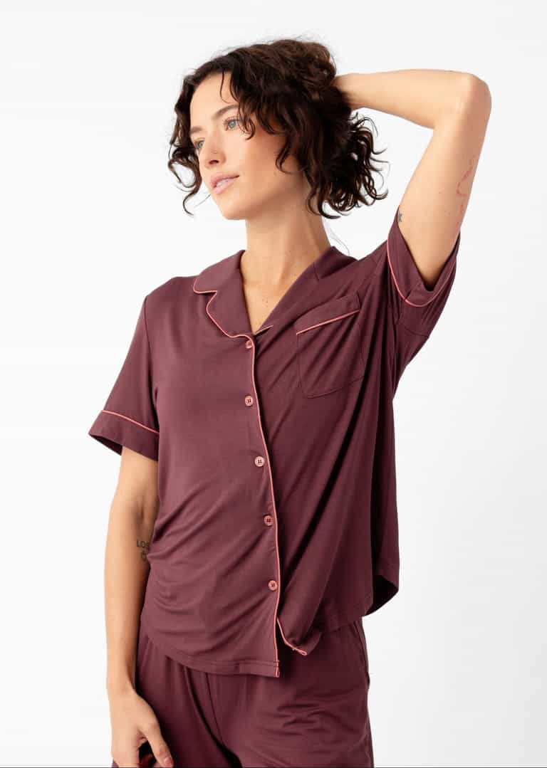 A woman models the Cozy Earth Women's Bamboo Stretch-Knit Short Sleeve Pajama Set, featuring a maroon, button-up top and matching shorts, standing in a relaxed pose against a plain white background.|Color:Burgundy