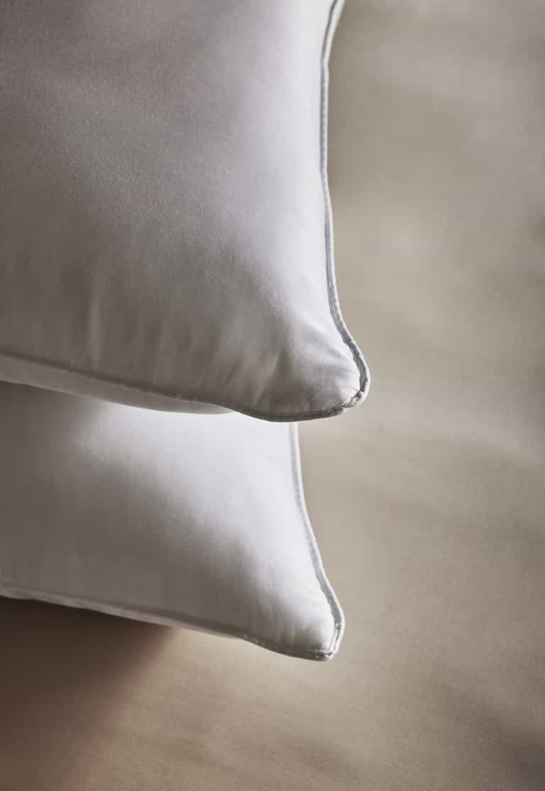 Two Bamboo Down Alternative Pillows by Cozy Earth with smooth, satiny pillowcases are neatly stacked on a light beige surface and viewed close up from the corner.Standard King