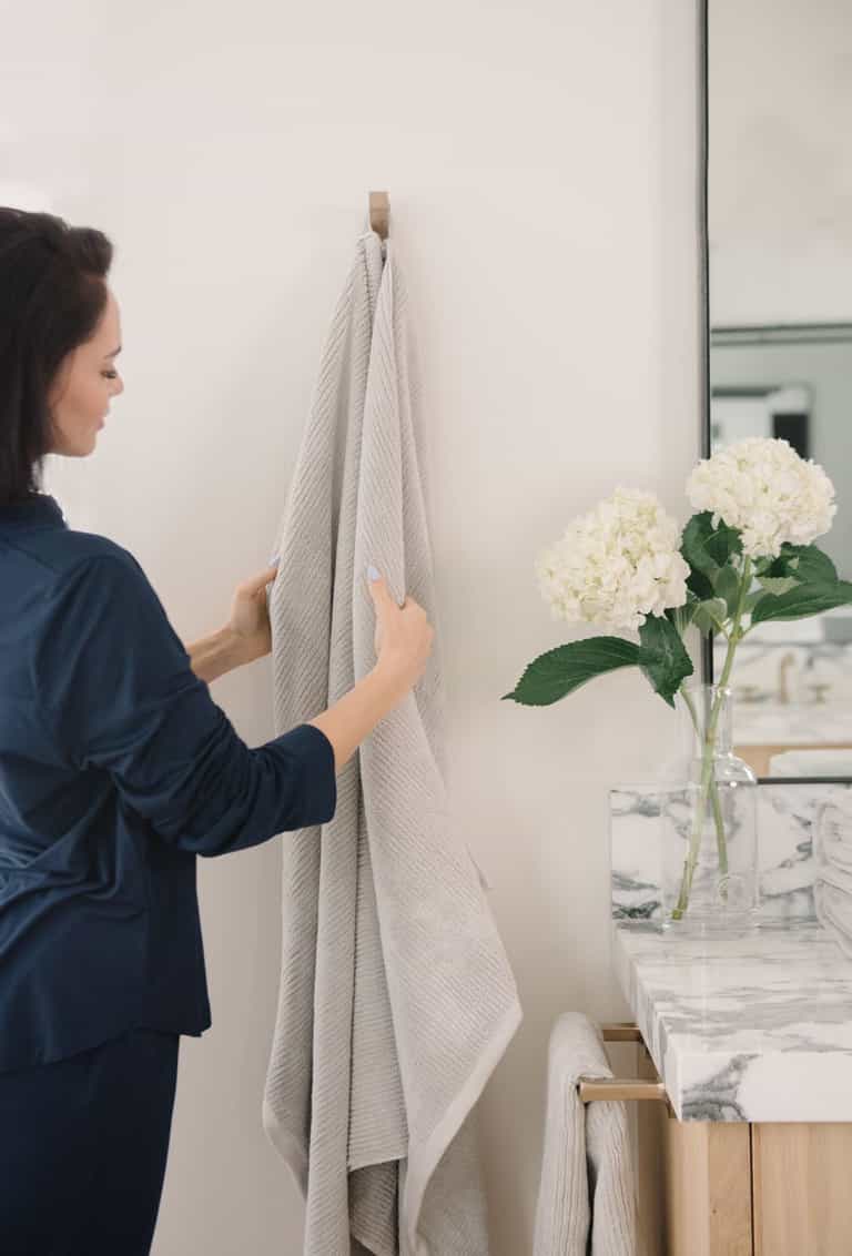 Ribbed Terry Bath Sheets in the color Light Grey. Photo of product taken in a bathroom next to a sink as a woman feels the towel. |Color:Light Grey
