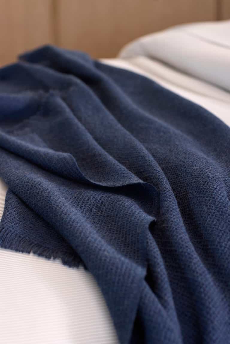 The Cozy Earth Alpaca Waffle Throw, a dark blue textured blanket with fringed edges, is draped casually over a neatly made bed with white sheets.|Color:Slate Blue
