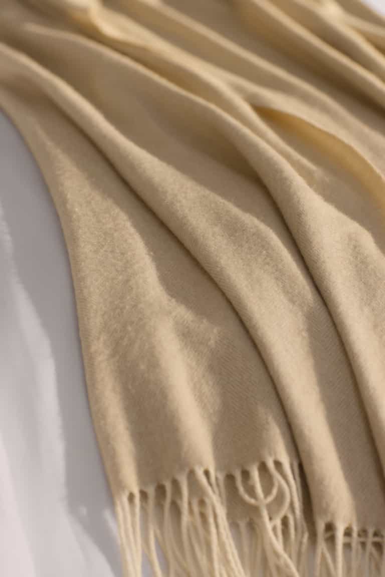 The Cozy Earth Bamboo Herringbone Tassel Throw, featuring a subtle herringbone texture and tasseled edges, is draped over a white surface in gentle folds.|Color:Oatmeal