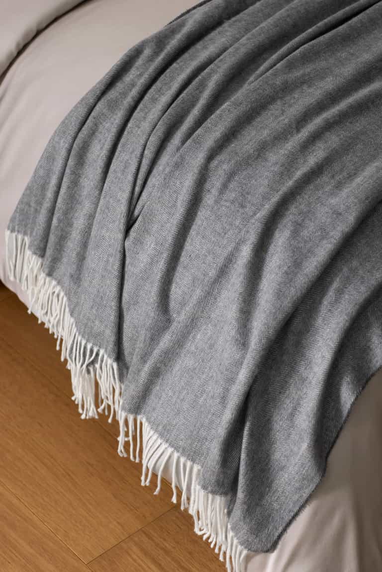 The Bamboo Herringbone Tassel Throw by Cozy Earth, featuring a gray herringbone pattern and white fringe, is draped over the edge of a neatly made bed on a wooden floor.|Color:Shadow