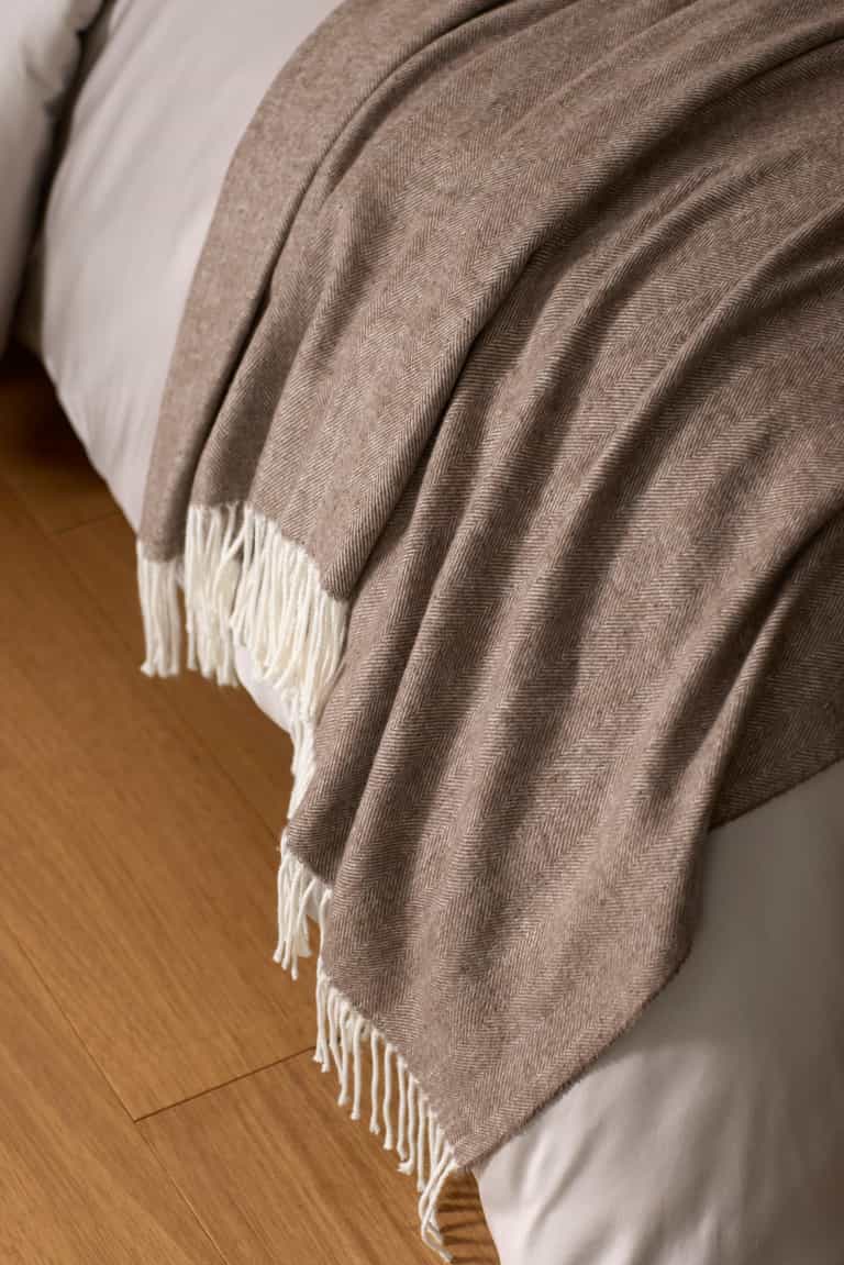 The Cozy Earth Bamboo Herringbone Tassel Throw, a soft light brown blanket with white fringe, is draped over a white bed resting on a wooden floor.|Color:Truffle