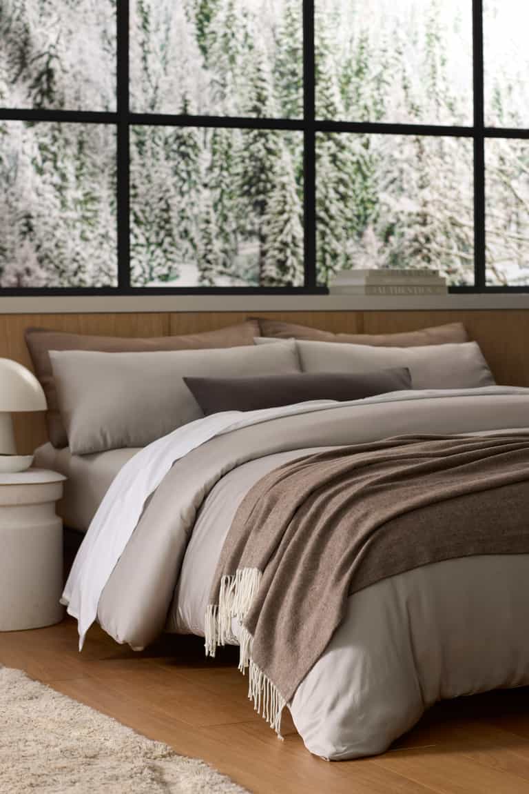 A cozy bed features Cozy Earth's Bamboo Herringbone Tassel Throw in beige and brown, set in a modern room with large windows framing a snowy forest view.|Color:Truffle