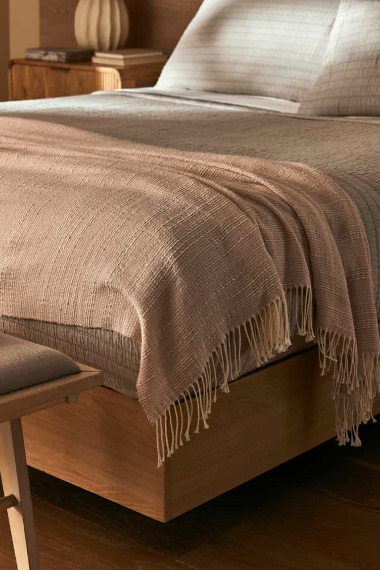 A close-up of a neatly made bed with the Cozy Earth Baja Tassel Throw draped at the foot. The wooden frame and neutral bedding create a cozy, minimalist vibe, complemented by the throw’s light texture.|Color:Himalayan Salt