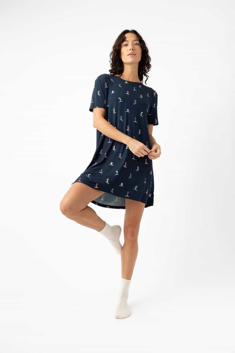 A woman stands on one leg against a white background, wearing the Cozy Earth Women's Bamboo Stretch-Knit Sleep Dress with small colorful prints and white socks. She looks slightly upward, holding the edge of her dress with one hand. |Color:Skiers Navy