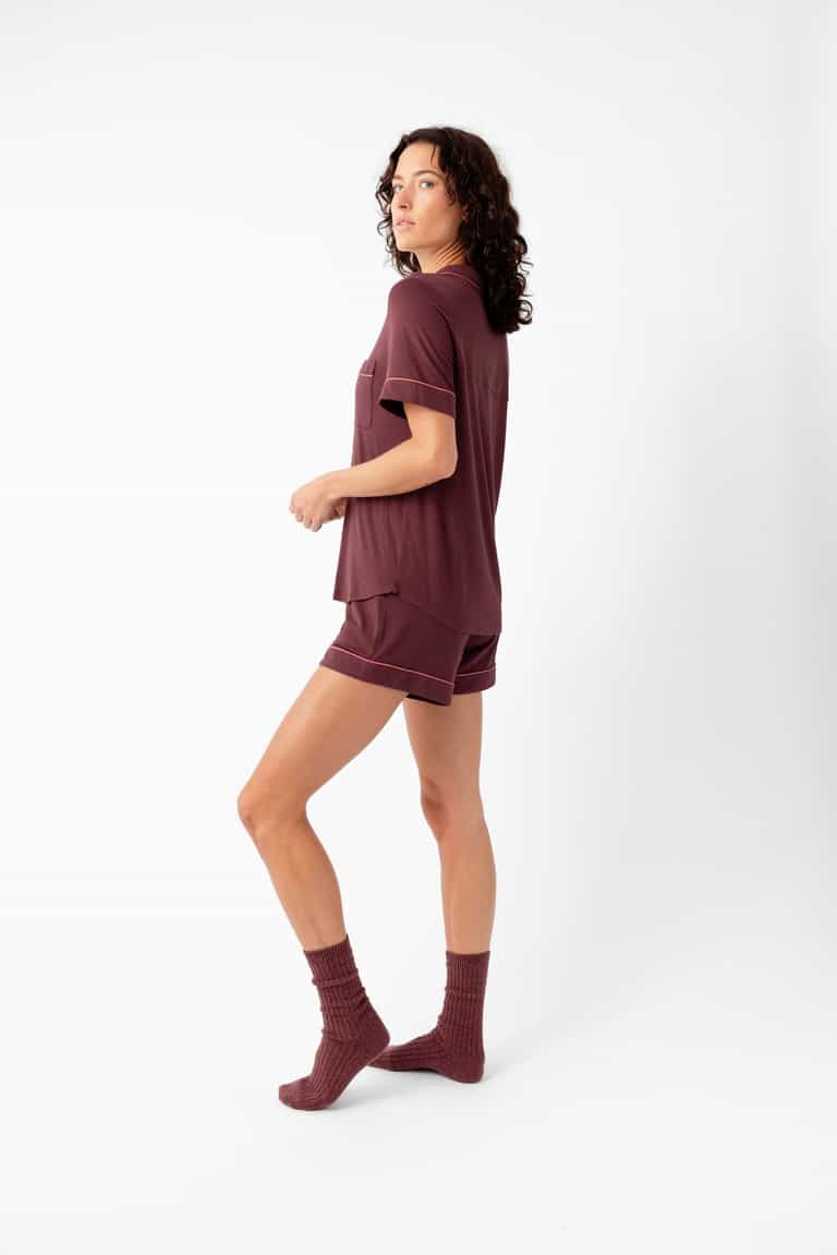 A woman with curly hair stands sideways in a Cozy Earth Women's Bamboo Stretch-Knit Short Sleeve Pajama Set—maroon shorts, short-sleeve top, and socks—against a white background.|Color:Burgundy