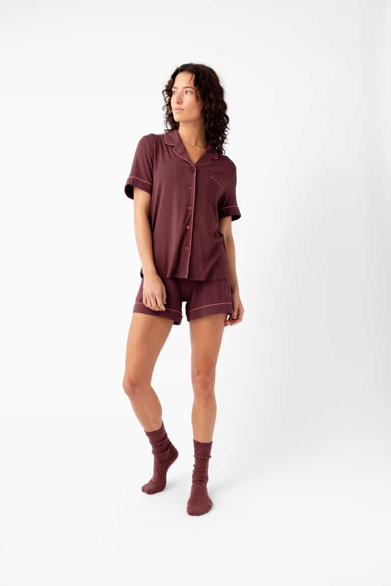 A woman with curly hair stands against a white background wearing Cozy Earth's Women's Bamboo Stretch-Knit Short Sleeve Pajama Set in burgundy with matching socks, looking to the side with a relaxed expression.|Color:Burgundy