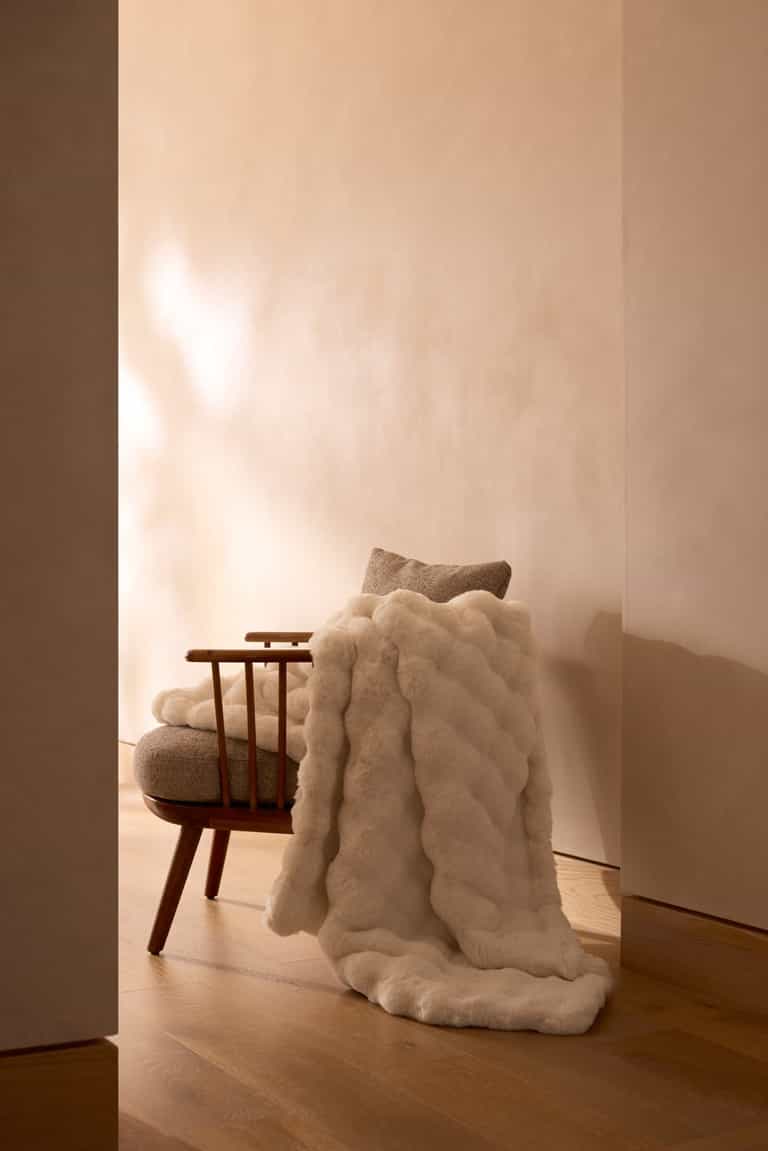 A wooden chair with a light cushion and a Cozy Earth Bubble Cuddle Blanket in plush white faux fur sits on a wooden floor against a beige wall in a softly lit, minimalist room. |Color:Creme