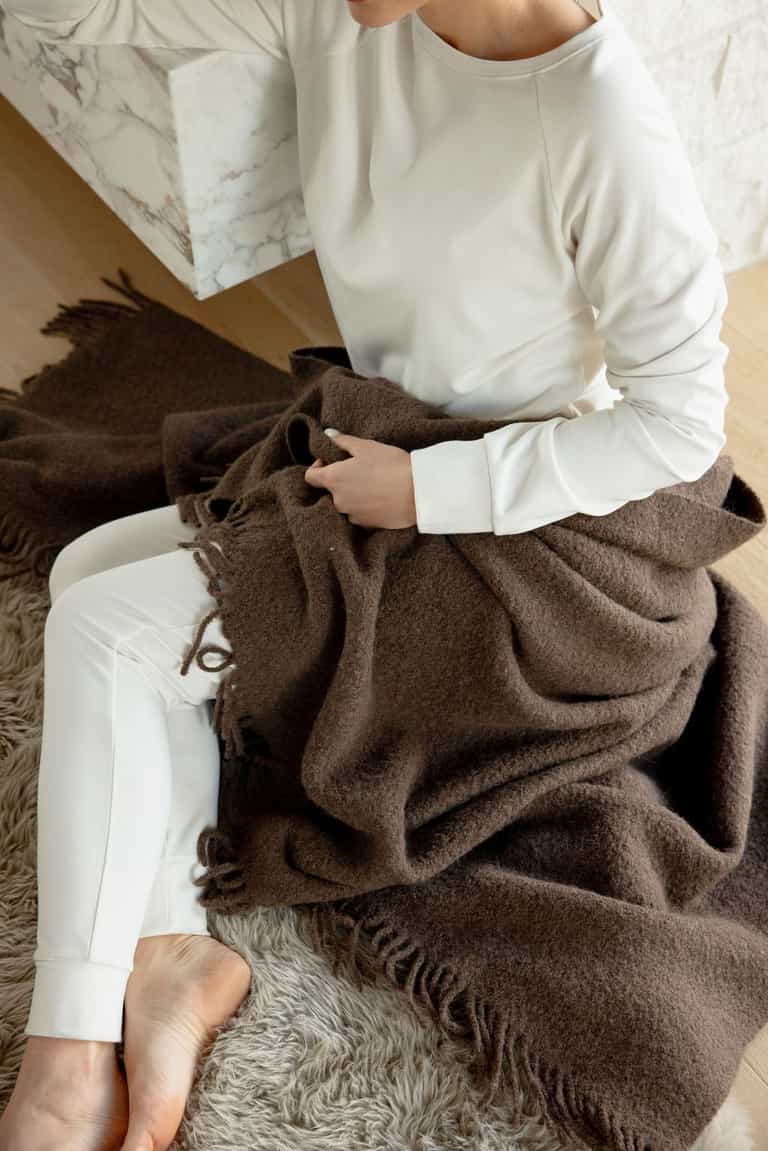 A person dressed in a white long-sleeve shirt and Cozy Earth Women's Bamboo Jogger Pants sits on a beige carpet with a brown blanket wrapped around their lap. Their feet are bare, and the person is sitting with one hand holding the blanket and the other resting on a marble surface. |Color:Ivory
