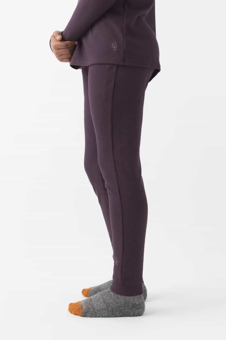 A child stands sideways in Cozy Earth Children's Bamboo Waffle Knit Base Layer Pant, paired with a matching purple thermal top and grey-orange socks, against a plain white background.|Color:Plum