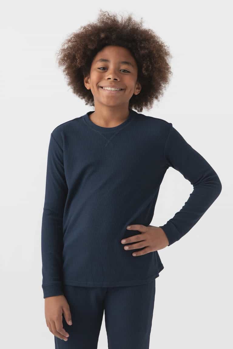 Smiling child with curly hair wears Cozy Earth's Children’s Bamboo Waffle Knit Base Layer Long Sleeve in navy blue, standing against a plain white background with one hand on their hip.|Color:Navy