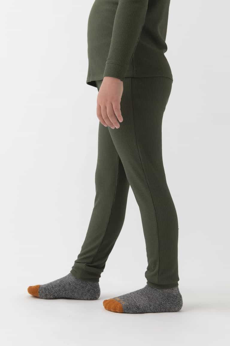 A child stands sideways on a white background, wearing Cozy Earth's Children's Bamboo Waffle Knit Base Layer Pant in green and a matching top. The child also has on gray socks with orange toes, and the face is not visible.|Color:Olive