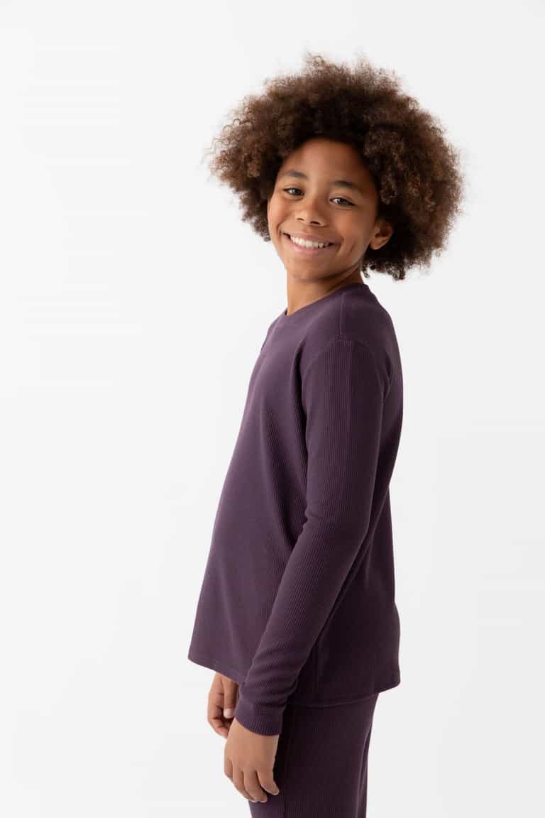 A smiling child with curly hair wears the Cozy Earth Children's Bamboo Waffle Knit Base Layer Long Sleeve in dark purple, paired with matching pants, standing against a plain white background and facing slightly to the side.|Color:Plum