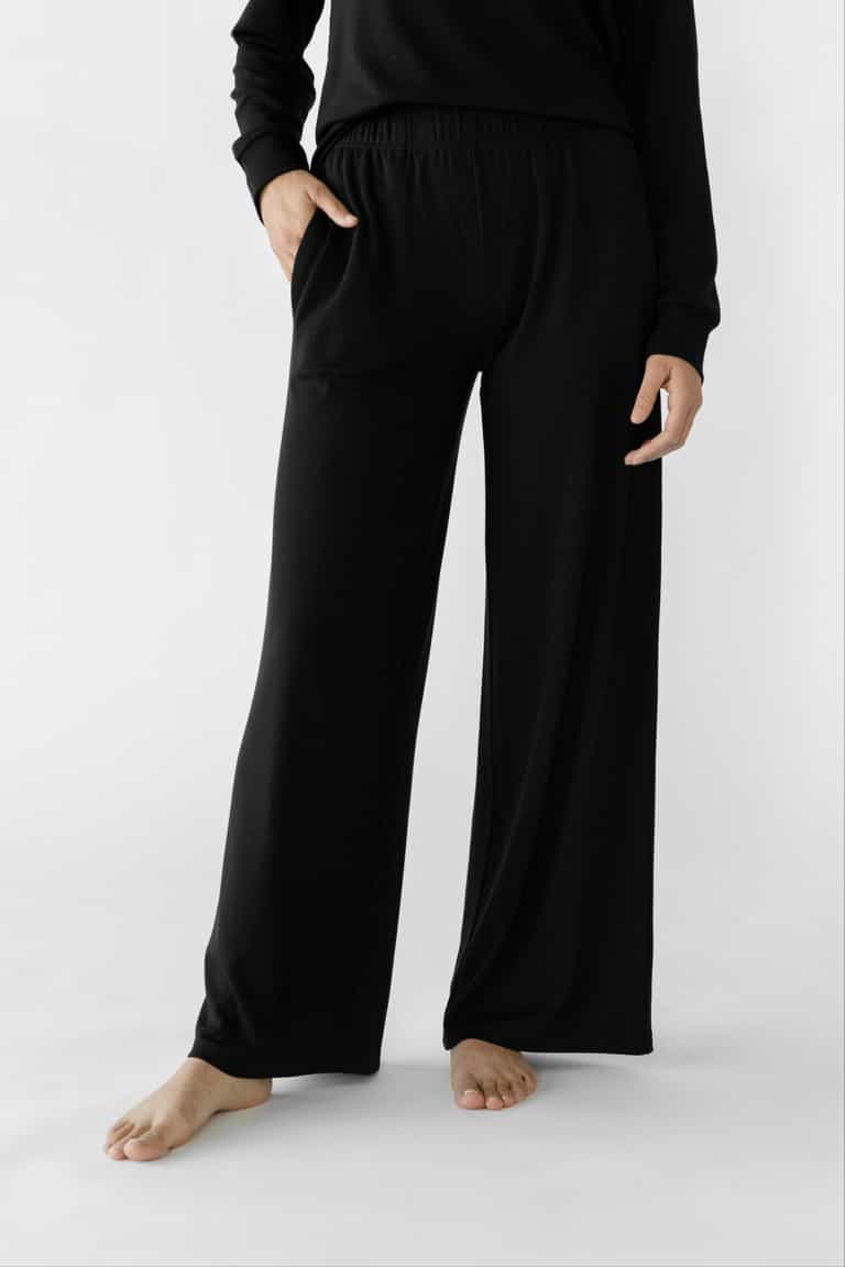 A person stands barefoot in a black Cozy Earth Women's Ultra-Soft Bamboo Wide Leg Pull On Pant & Pullover Set against a plain white background, partially showing the upper body with one hand resting in a pocket.|Color:Black
