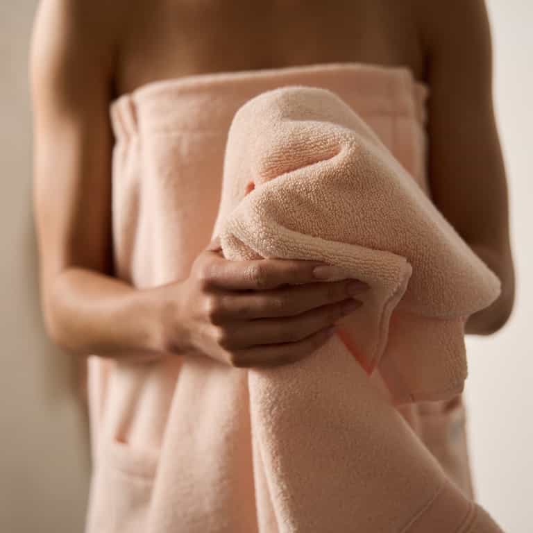 A person wrapped in a soft Luxe Bath Towel in peach from Cozy Earth holds another towel, evoking a calm and serene atmosphere against a neutral background. |Color:Blossom