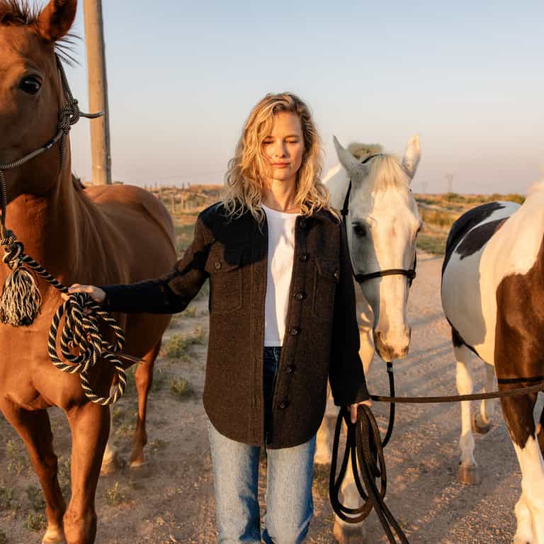 A woman with long, curly blonde hair stands outdoors holding the reins of three horses. She is wearing a Cozy Earth Women's Boucle Shacket over a white shirt and blue jeans. The sun is low on the horizon, casting a warm glow over the scene. The background includes a dirt path and a fence. |Color:Coal