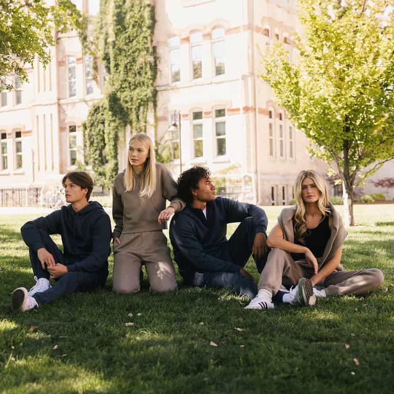 Four people sit on the grass outdoors, all dressed in casual and comfortable attire. Among them, one individual wears the Men's CityScape Sweatpant from Cozy Earth. In the background, there's a large light-colored building with windows and trees, suggesting the scene is set in a park or university campus. |Color:Eclipse