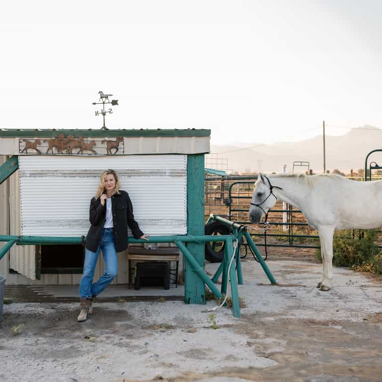 A person wearing a Cozy Earth Women's Boucle Shacket and blue jeans stands outdoors next to a small structure with a weather vane on top, featuring horse decorations. A white horse is tethered to a post near the person. The background includes fencing and distant mountains. |Color:Coal