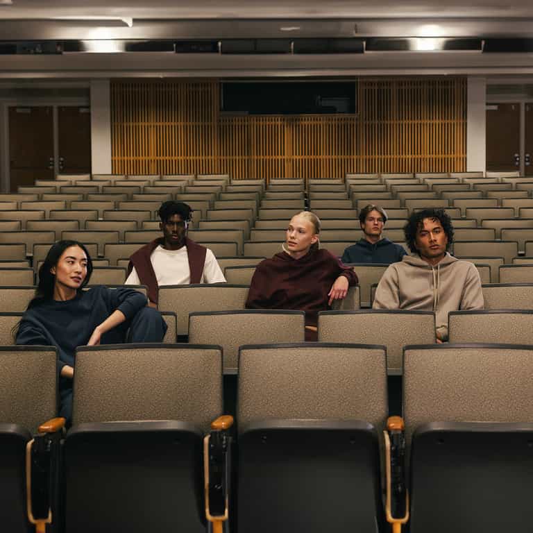 Five people are sitting spread out in an empty lecture hall with gray seats and light wood paneling on the back wall. They are casually dressed, some of them wearing the Women's CityScape Crewneck from Cozy Earth, each looking in different directions and giving the scene a contemplative atmosphere. |Color:Eclipse