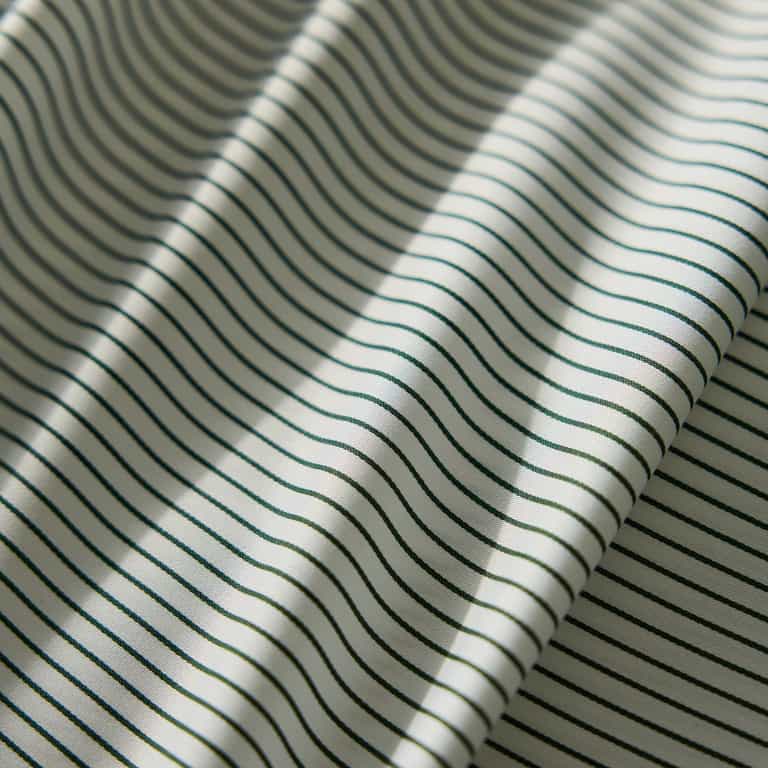 Close-up of the Bamboo Flat Sheet from Cozy Earth, featuring a cream-colored fabric with thin, evenly spaced black stripes. The sheet lies flat with gentle folds, creating soft shadows and highlights. |Color:Jade Pinstripe