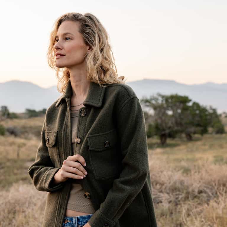 A woman with wavy blonde hair stands outdoors in a grassy landscape with distant mountains. She wears a dark green Cozy Earth Women's Boucle Shacket over a beige top and gazes to the left, her hand near the jacket's buttons. The sky is pale, and the scene has a serene, natural ambiance. |Color:Olive