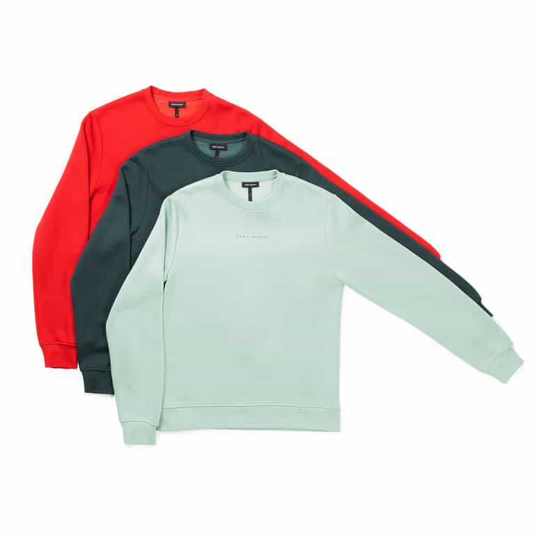The display features three Men's CityScape Crewneck sweatshirts from Cozy Earth, each showcased in light green, dark green, and red. Their simple design is free from visible patterns, with sleeves extended to the side against a white background. |Color:Wintergreen