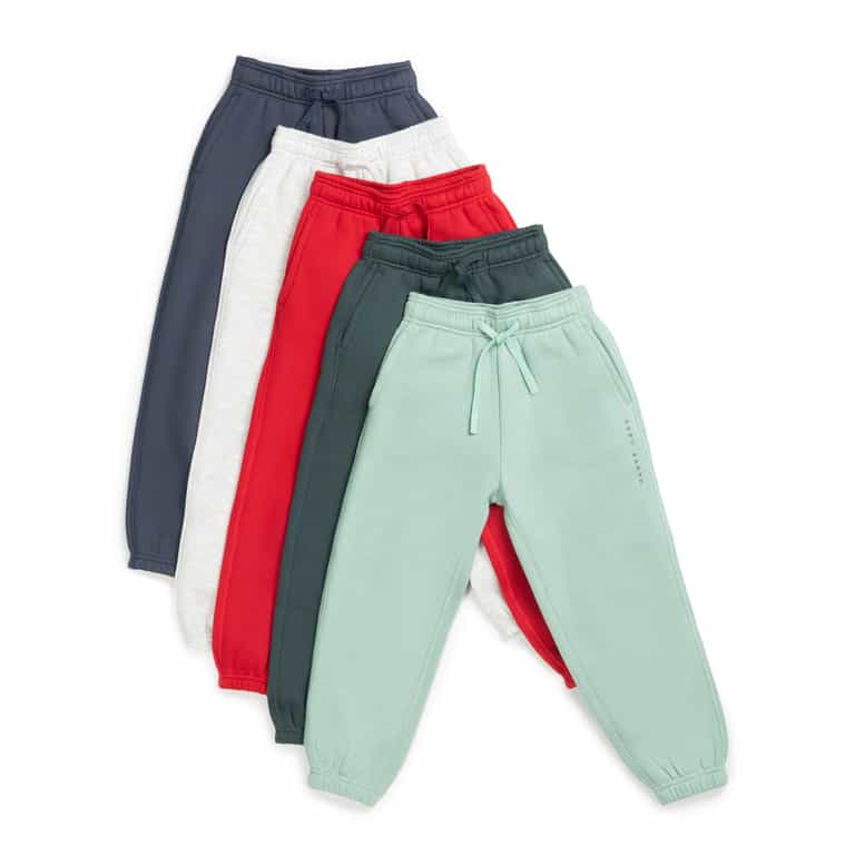 A set of five Kid's CityScape Sweatpants from Cozy Earth, featuring an array of colors like dark blue, light gray, red, dark green, and mint green, is laid out in a staggered stack. Each sweatpant includes an elastic waistband and drawstrings. |Color:Spruce