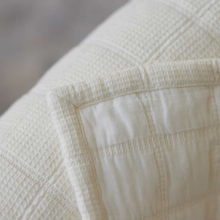 Close-up of the Waffle Windowpane Blanket by Cozy Earth. This cream-colored blanket showcases a textured fabric with a checkerboard pattern and subtle grid design. The meticulous stitching detail along the edges gives it a neat and tailored appearance. |Color:Creme