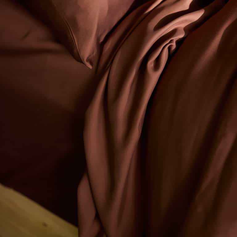 Close-up of a bed featuring Cozy Earth's Bamboo Flat Sheet in deep brown, with a slightly wrinkled, rich brown comforter cascading over the edge. The fabrics have a smooth, silky texture and exude a sense of warmth and coziness. |Color:Terra