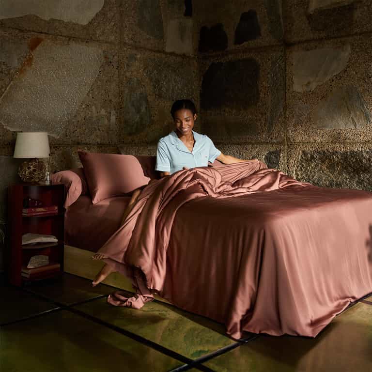 A person smiles and holds up the Bamboo Flat Sheet - Wholesale by Cozy Earth while sitting on a bed adorned with peach-colored sheets and pillows. The room features a rugged stone wall, with a nightstand that has a lamp and a book beside the bed. The person is dressed in light blue pajamas, and the floor has a reflective surface. |Color:Terra