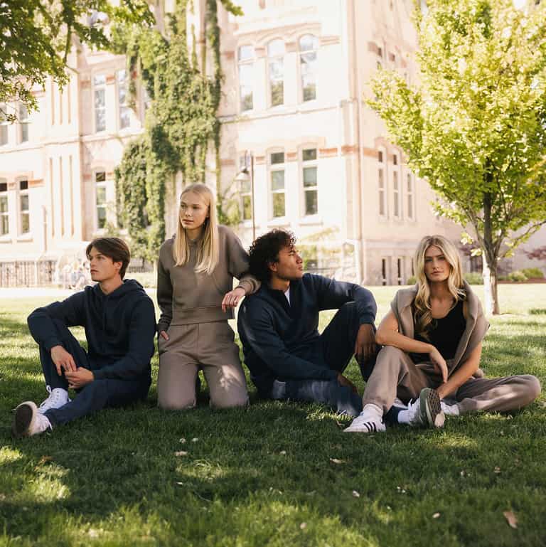 Four people, two men and two women, are sitting on grass in front of a building with large windows and walls covered in ivy. They are dressed in casual athletic wear, with the men sporting the Men's CityScape Quarter Zip from Cozy Earth. The scene suggests a relaxed and friendly atmosphere on a sunny day. |Color:Eclipse
