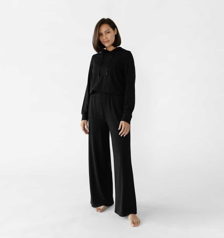 A woman stands in a relaxed pose wearing a black loungewear set from Cozy Earth, featuring the Women's Ultra-Soft Bamboo Wide Leg Pull On Pant TALL and a matching hooded sweatshirt. |Color:Black