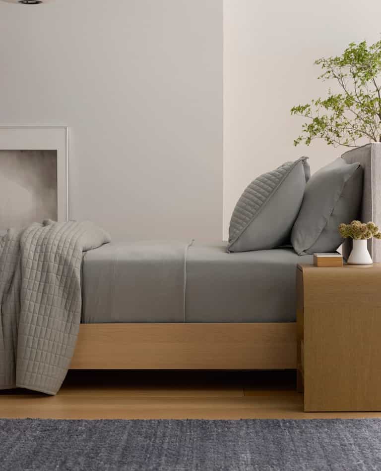 A neatly made bed features the Cozy Earth Bamboo Jersey Flat Sheet in gray, paired with pillows. Next to it, a wooden nightstand holds a potted plant, books, and a vase, while a hanging light and minimalist decor create a calm, modern bedroom.|Color:Stone