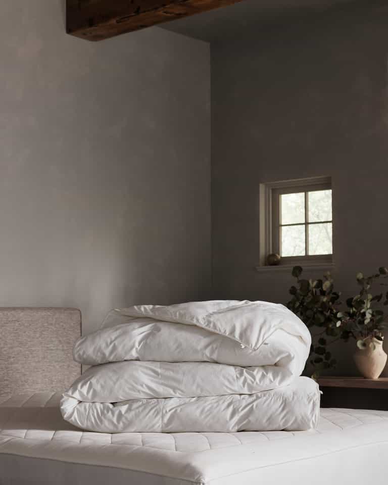 A neatly folded Cozy Earth Down Alternative Comforter rests on a bed in a softly lit, minimalist bedroom with gray walls, a small window, and a potted plant on a wooden table.Coal Stripe/Striped Alabaster, Laurel Stripe/Striped Bone