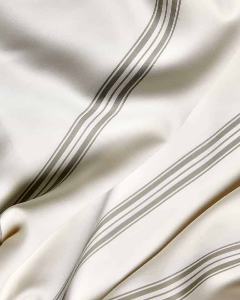 A close-up of Cozy Earth’s Bamboo Sheet Set shows ivory fabric with parallel gray stripes, soft folds, and a smooth texture.|Color:Baja Stripe in Laurel