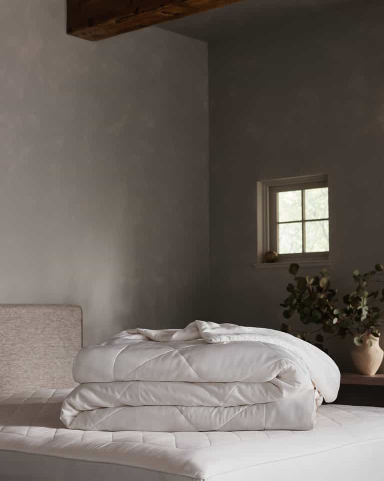 A neatly folded Cozy Earth Bamboo Viscose Comforter rests on a mattress in a softly lit bedroom with gray walls, a small window, and a vase with greenery atop a wooden nightstand. Charcoal, Driftwood, Oat, Creme, White, Dove Grey, Light Grey, Shore, Pacific Blue, Peony, Walnut
