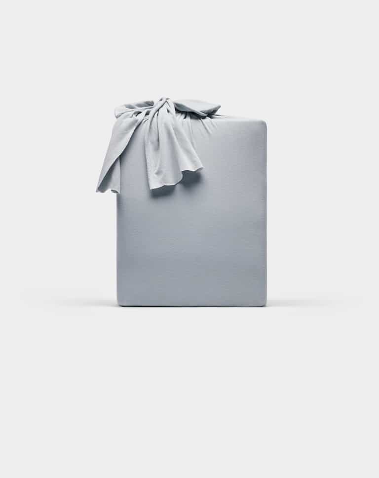 A rectangular Bamboo Jersey Sheet Set by Cozy Earth, wrapped in smooth, light gray fabric with a knot on top, displayed against a plain white background.|Color:Shore