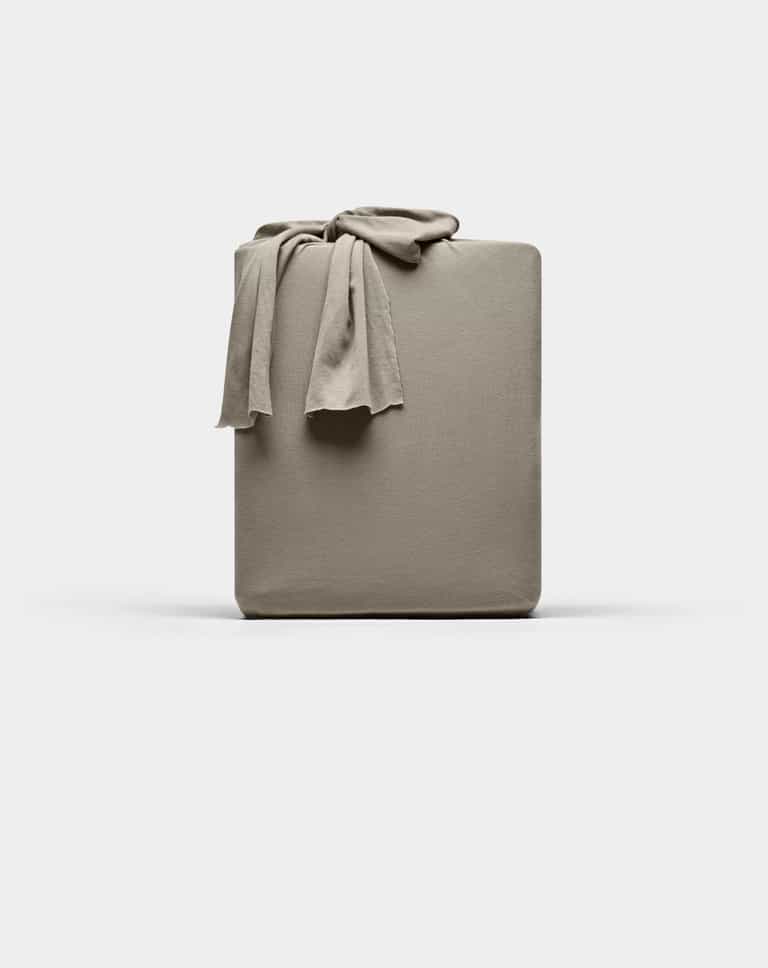 A taupe Cozy Earth Bamboo Jersey fitted sheet is neatly folded with one corner draped over the top, displayed against a plain white background.|Color:Stone