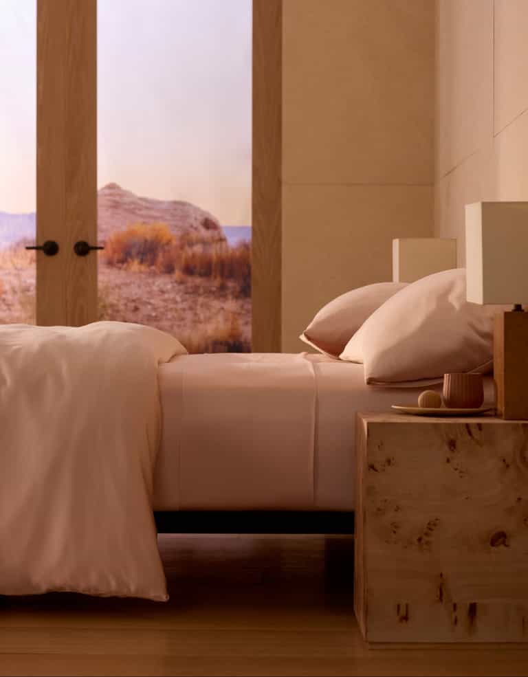 A neatly made bed with Cozy Earth's Bamboo Sheet Set in light pink sits next to a wooden nightstand with a lamp. Large windows showcase a scenic sunlit desert, adding to the minimalist, cozy atmosphere. |Color:Peony