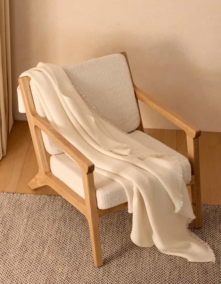 A modern wooden armchair with light beige upholstery sits on a herringbone rug, draped with the Cozy Earth Alpaca Waffle Throw. The soft blanket and minimal setting create a warm, inviting atmosphere.|Color:Creme