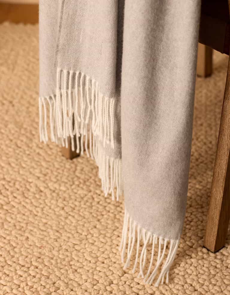 The Bamboo Herringbone Tassel Throw by Cozy Earth, in light gray, is draped over a wooden chair with a textured beige rug placed on the floor beneath.|Color:Cloud
