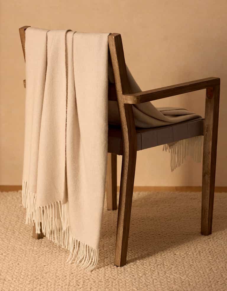 The Cozy Earth Bamboo Herringbone Tassel Throw in beige is draped over a wooden chair with a cushioned seat, set atop a textured cream carpet against a neutral wall.|Color:Oatmeal